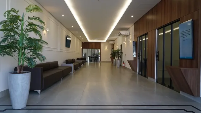 Corredor de acesso às capelas, com poltronas encostadas na parede, vaso com planta e iluminação de sanca