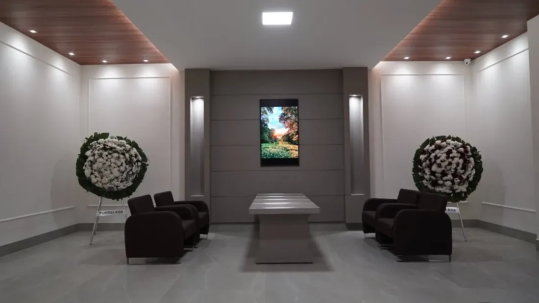 Sala de velório com duas coroas de flores em cavaletes, poltronas escuras, mesa de centro e forro de madeira